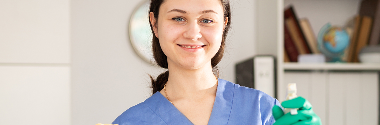 Mulher branca sorrindo utilizando luvas verdes e uniforme de limpeza azul